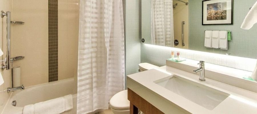 A modern bathroom with a bathtub, white shower curtain, toilet, sink with a large mirror above, a towel rack with white towels, and a framed picture on the wall—reminiscent of the luxurious amenities you might find in hotels near West Edmonton Mall.