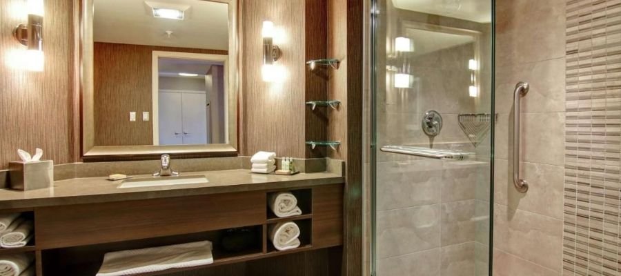 Bathroom with a large mirror above a sink vanity, shelves with rolled towels, and a glass-enclosed walk-in shower with gray tiles and a grab bar, perfect for guests staying at hotels near West Edmonton Mall.