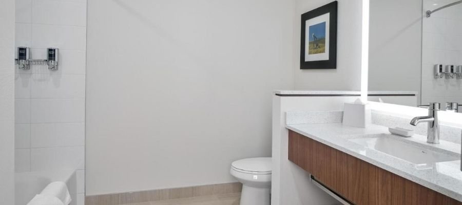 A modern bathroom near hotels near West Edmonton Mall features white walls, a toilet, a sink with a wooden cabinet, a wall-mounted faucet, a mirror with built-in lighting, a towel rack, and a framed picture.