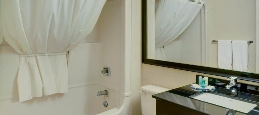 A bathroom in the hotels near West Edmonton Mall features a white shower curtain, shower-tub combo, black countertop, sink, mirror, and towel rack with white towels. Various toiletries are placed on the countertop.