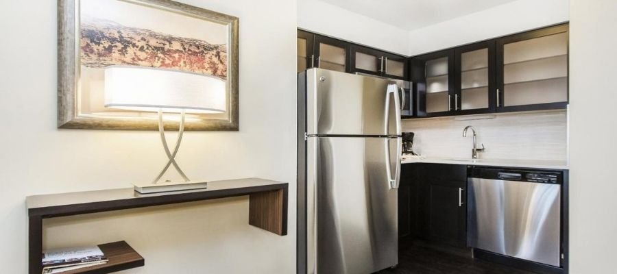 Compact kitchen with stainless steel appliances, including a fridge and dishwasher. Dark cabinets contrast with the white backsplash. Adjacent area features a wall-mounted shelf and framed artwork, perfect for those seeking hotels near West Edmonton Mall.