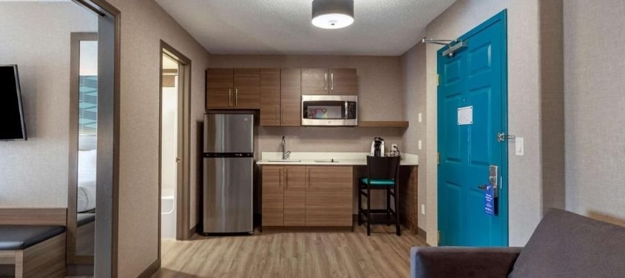 A small apartment kitchenette with wood cabinets, stainless steel fridge, microwave, sink, a teal door, and light hardwood floor. A barstool and partial views of adjoining spaces are visible—reminiscent of the cozy amenities found in hotels near West Edmonton Mall.