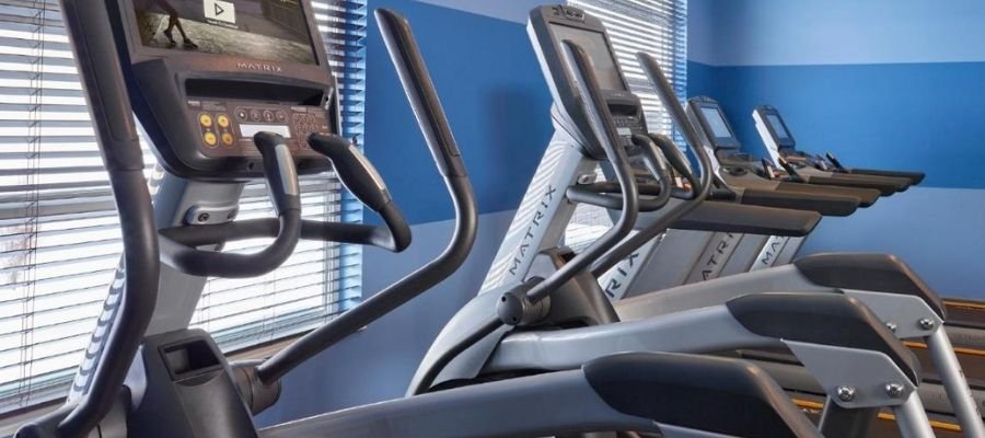 Several elliptical machines with digital displays are lined up in a gym with blue walls and large windows, providing an invigorating workout space for guests staying at hotels near West Edmonton Mall.