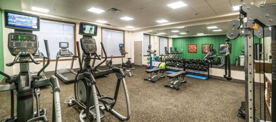 A fitness center with various exercise machines including treadmills, ellipticals, weight benches, and dumbbells. Mirrors cover one wall, reflecting the equipment. TVs are mounted above the machines—perfect for guests staying at hotels near West Edmonton Mall looking to stay active.
