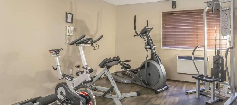A small gym room with a stationary bike, an elliptical machine, a weight bench, and a multi-station weight machine. The room has beige walls, wooden flooring, and a window with blinds. Perfect for guests seeking hotels near West Edmonton Mall who want to stay active during their visit.