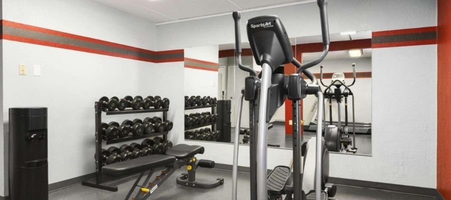 A gym room with exercise equipment, including an elliptical machine, a weight bench with dumbbells, and a large wall mirror. The walls are white with a red and grey stripe. Perfect for guests at hotels near West Edmonton Mall looking to maintain their fitness routine.