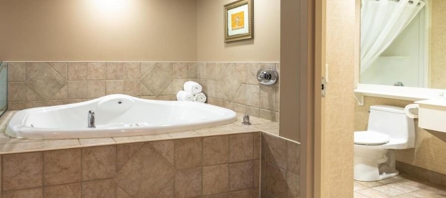 A bathroom in hotels near West Edmonton Mall features a large corner bathtub surrounded by beige tiles, rolled white towels on the ledge, a framed picture on the wall, and a white sink with a mirror, shower curtain, and toilet.