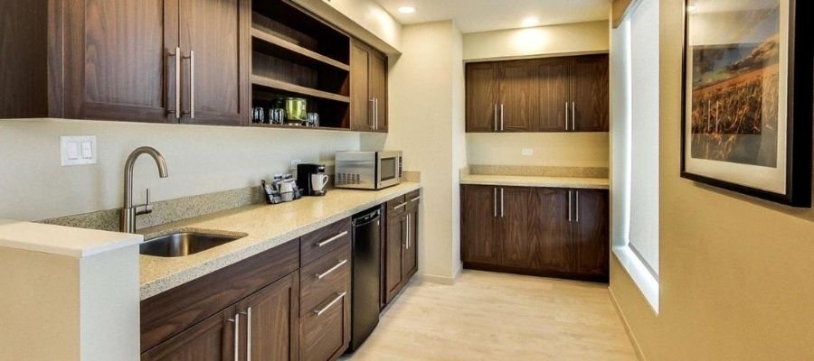 A small kitchen with wooden cabinetry, a sink, a dishwasher, a microwave, and ample counter space. A picture frame is mounted on the right wall, and under-cabinet lighting illuminates the workspace. Perfect for travelers seeking hotels near West Edmonton Mall who enjoy self-catering options.