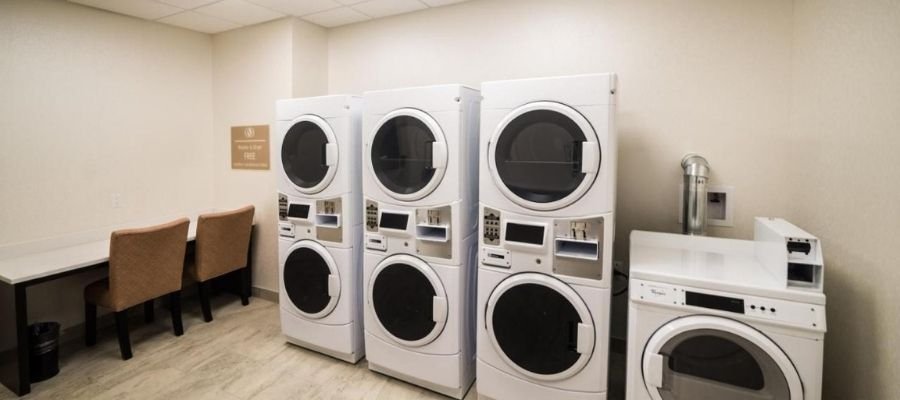A laundry room with three stacked washer-dryer units, one standalone dryer, and a table with two chairs against the wall, similar to the convenient amenities found in hotels near West Edmonton Mall.