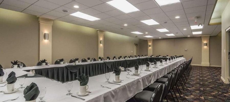 A long, U-shaped conference table set for a formal meeting in a well-lit room with gray walls and ceiling lights. Cups, glasses, cutlery, and napkins are neatly arranged at each place setting—perfect for events hosted by hotels near West Edmonton Mall.