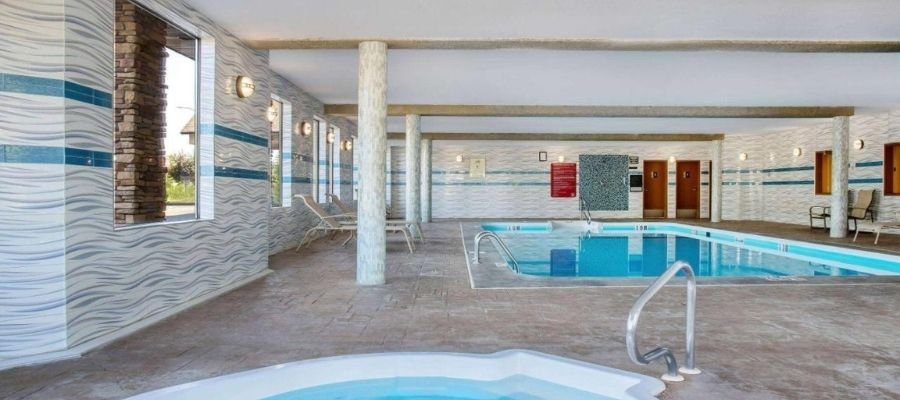 Indoor swimming pool area with adjacent hot tub in hotels near West Edmonton Mall. There are lounge chairs along the tiled walls and large windows allowing natural light into the space.