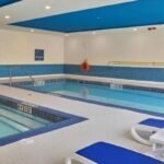 Indoor swimming pool area with a separate hot tub, blue lounge chairs, and pool safety signage. The walls and flooring are tiled in white and blue. Lifebuoy and clock visible on the wall, reminiscent of those found in the finest hotels near West Edmonton Mall.