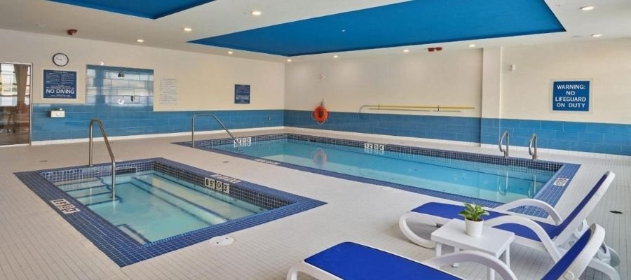 Indoor pool area with a main swimming pool and a hot tub. The space is brightly lit and features blue and white decor. Two lounge chairs with blue cushions are positioned near the pools, perfect for guests exploring hotels near West Edmonton Mall.