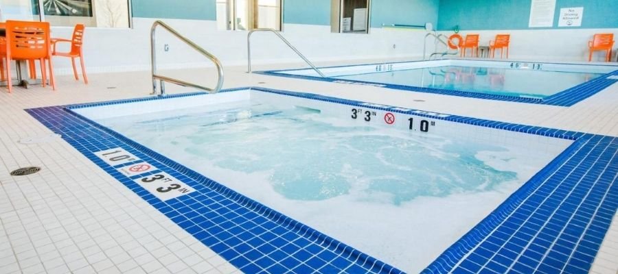 Indoor pool area with a small hot tub in the foreground and a larger pool in the background. Several orange chairs and a table are situated near the pool, reminiscent of amenities you might find at hotels near West Edmonton Mall.