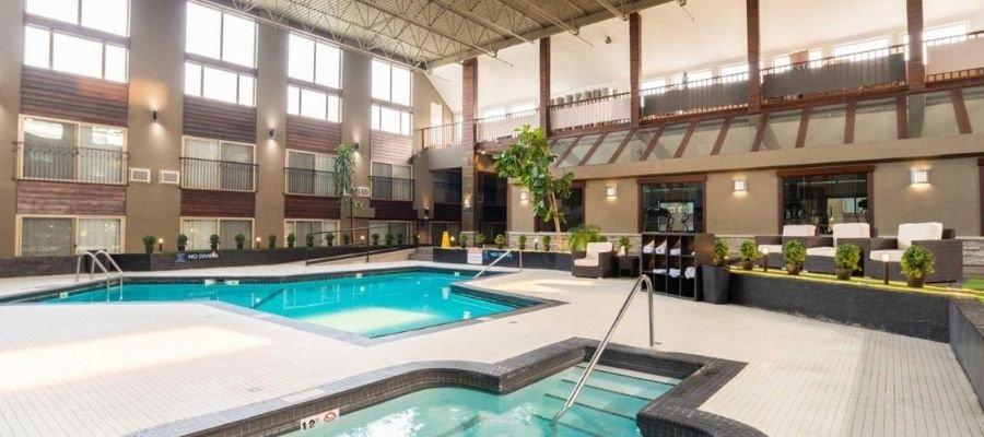 Indoor hotel pool area featuring a large swimming pool and a smaller hot tub in the foreground. The space includes greenery, comfortable seating, and large windows for natural light. Hotels near West Edmonton Mall offer such luxurious amenities for a relaxing stay.