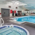 Indoor swimming pool area with a hot tub in the foreground, lounge chairs, and tables around the pool. Bright lighting with white and gray decor, large windows on the right side allow natural light in. Perfect for guests looking for hotels near West Edmonton Mall.