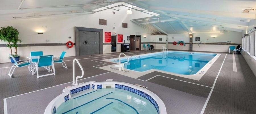 Indoor swimming pool area with a hot tub in the foreground, lounge chairs, and tables around the pool. Bright lighting with white and gray decor, large windows on the right side allow natural light in. Perfect for guests looking for hotels near West Edmonton Mall.