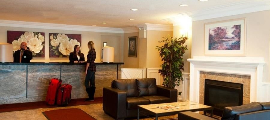 A hotel reception area with a front desk, a receptionist assisting a guest, two suitcases, sofas, a coffee table, a plant, and wall art, exudes warmth akin to the cozy lodgings found in hotels near West Edmonton Mall.