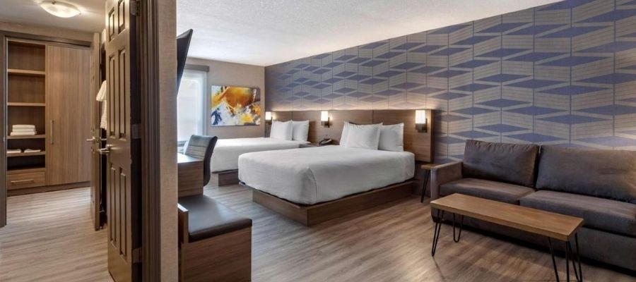 A modern hotel room near West Edmonton Mall features two double beds, a blue geometric-patterned accent wall, a desk, a sofa, and a wooden coffee table. The flooring is hardwood, and an open closet area is visible.