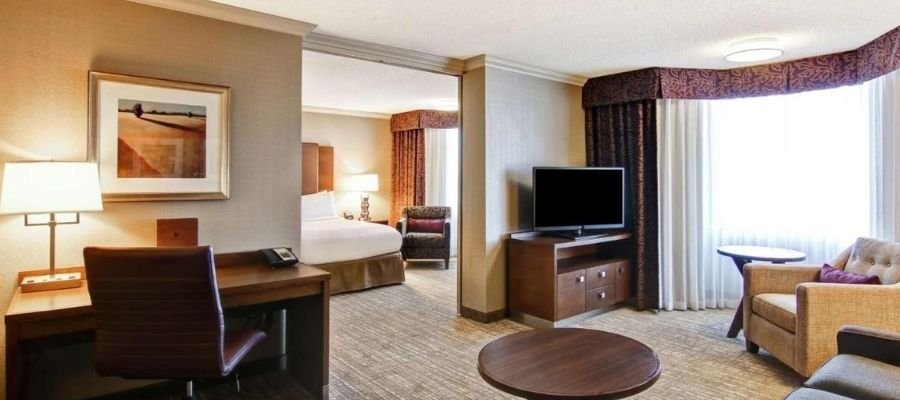 Spacious hotel room with a king-sized bed, desk, TV, armchair, coffee table, and a large window with curtains. The wall is adorned with a framed artwork. Perfect for travelers seeking hotels near West Edmonton Mall.