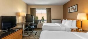 Hotel room with two double beds, a flat-screen TV, a writing desk with an office chair, a lamp, and a window with curtains. The walls are painted in neutral tones, and a framed picture hangs above the beds. Ideal for families visiting hotels near West Edmonton Mall.