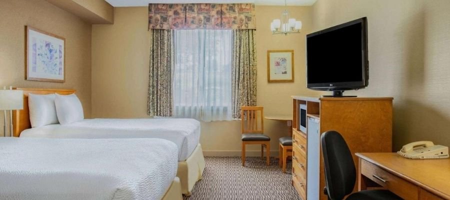 A hotel room with two white-sheeted beds, a television on a dresser, a desk with a chair, a window with curtains, and framed artwork on the walls is just what you can expect from hotels near West Edmonton Mall.