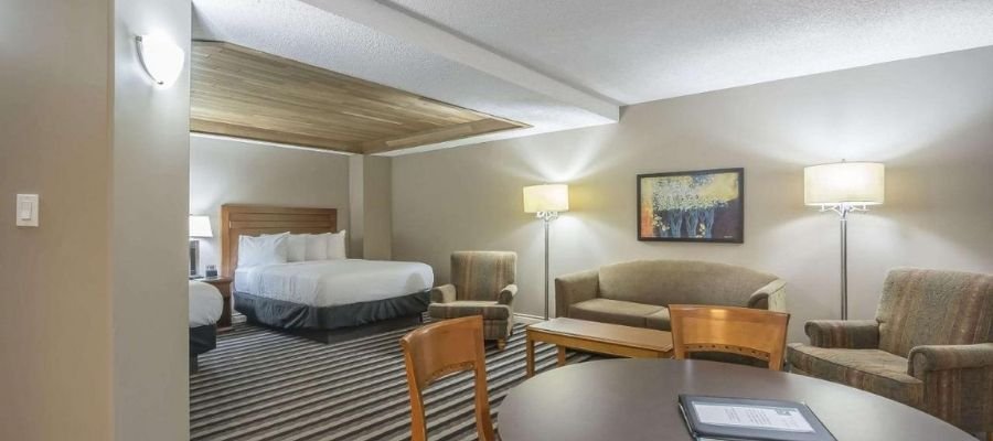 Hotel room with a king-sized bed, two armchairs, a sofa, and a round table. Room features modern lighting, a neutral color scheme, and a striped carpet. Decor includes a framed painting on the wall. Ideal for those seeking hotels near West Edmonton Mall.
