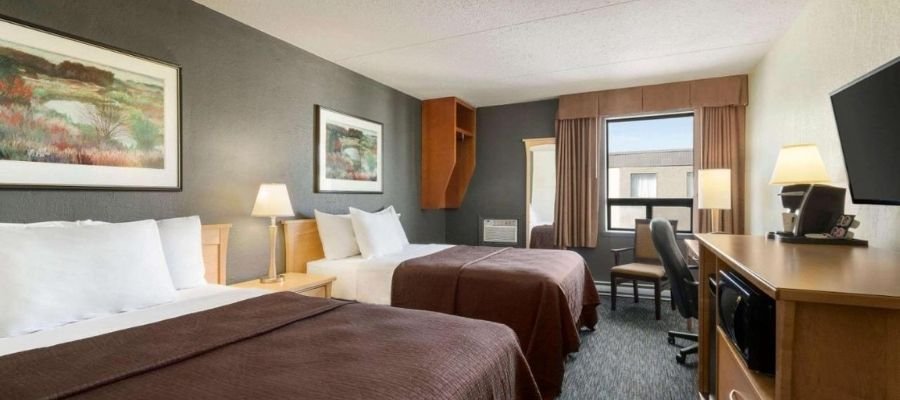Hotel room with two double beds, dark brown bedspreads, wooden furniture, a window with closed curtains, a mirror, and framed landscape art on the walls. Perfect for visitors seeking hotels near West Edmonton Mall.