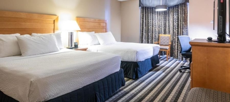 Hotel room with two double beds, a flat-screen TV, a desk with a chair, and a window with patterned curtains. Located among hotels near West Edmonton Mall, this room features neutral tones and striped carpet flooring.