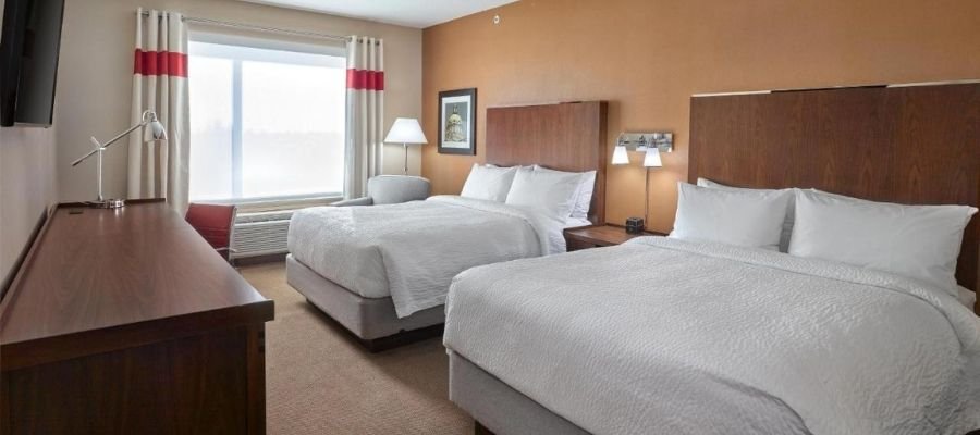 A hotel room near West Edmonton Mall features two double beds, wooden headboards, white bedding, a desk with a lamp, a chair by the window, and neutral-colored walls.