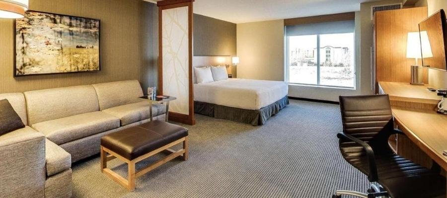 A modern hotel room near West Edmonton Mall featuring a king-sized bed, a grey sofa, a wooden coffee table, a desk with a chair, and a large window. The room also includes a wall painting and carpeted floor for added comfort.