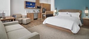 Modern hotel room featuring a large bed, a sofa, a kitchenette with microwave and sink, a small dining table, and wood furnishings. The accent wall is painted blue. Perfect for those seeking hotels near West Edmonton Mall.