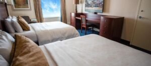 A hotel room with two double beds, wood furniture, a desk and chair, a wall-mounted TV, and a large window with a blue sky view awaits you at one of the fine hotels near West Edmonton Mall.