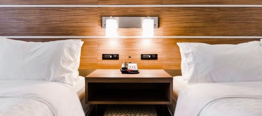 A modern hotel room, perfect for those visiting hotels near West Edmonton Mall, features two beds with white linens and pillows. A wooden headboard with built-in lights and a nightstand holding a phone are positioned between the beds.