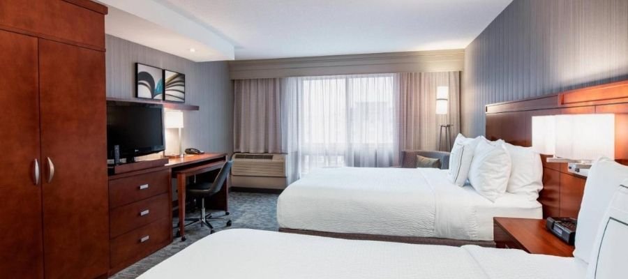 A hotel room with two double beds, a wooden desk with a chair, a TV on a wooden stand, a window with curtains, and modern decor, conveniently located near West Edmonton Mall.
