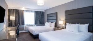 A hotel room near West Edmonton Mall features two neatly made double beds, gray upholstered headboards, a desk, a lamp, a flat-screen TV, and a window with curtains.