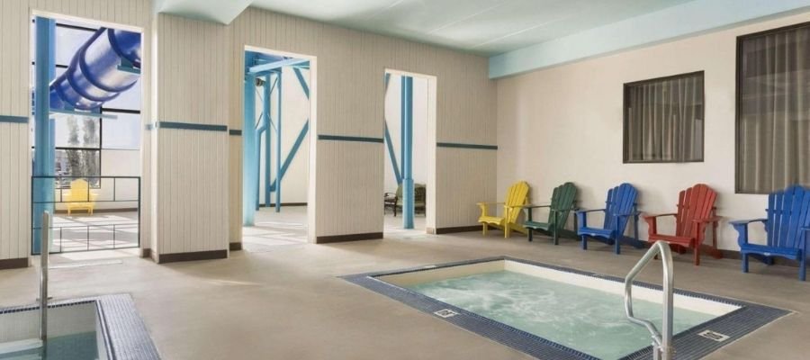 Indoor pool area with a small whirlpool spa surrounded by colorful Adirondack chairs and large windows in the background. A blue water slide is partially visible through an open wall. Perfect for families staying at hotels near West Edmonton Mall.