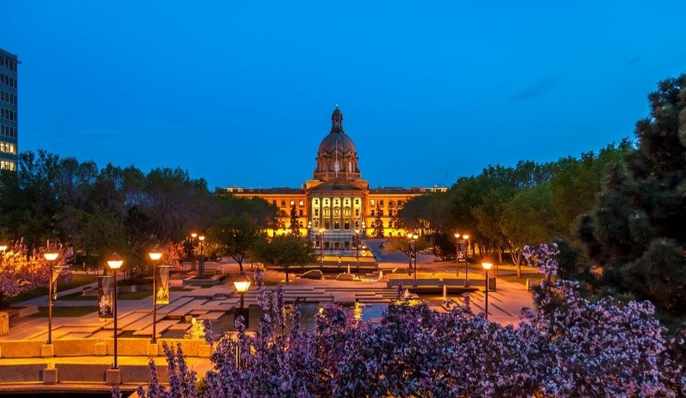 View of an illuminated domed building surrounded by trees and flower beds at dusk, with a well-lit plaza and walking paths in the foreground. This picturesque scene is one of the many attractions in Edmonton Alberta that captivate visitors.