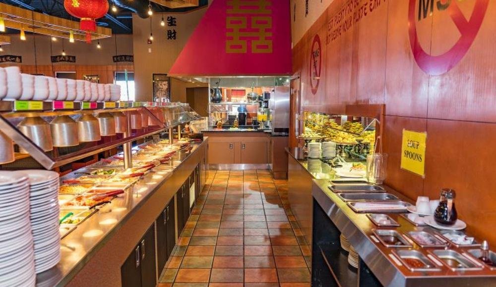 The interior of a buffet restaurant in Edmonton features food trays on long counters, stacks of plates, and signs indicating various food categories.
