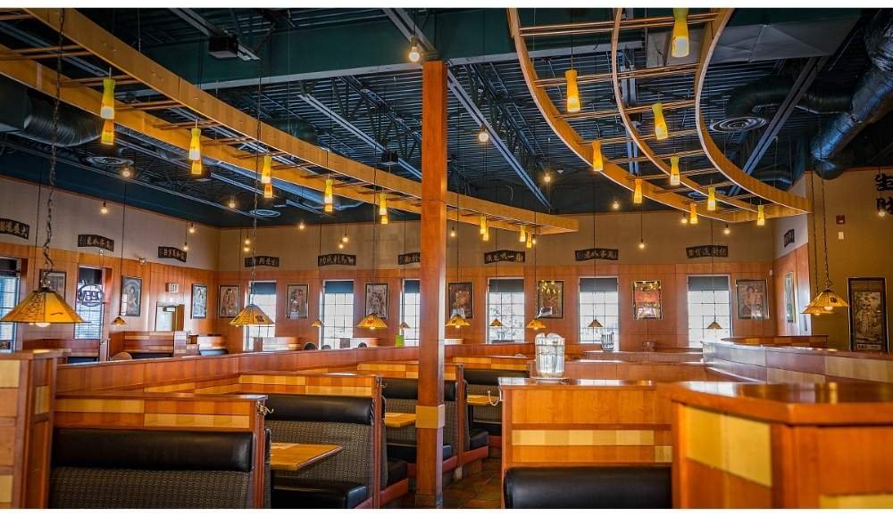 An empty restaurant with wooden booths, pendant lights, and decorative ceiling elements offers a cozy ambiance. Walls adorned with framed artwork and windows add a touch of elegance, setting the perfect scene for delightful buffets in Edmonton.