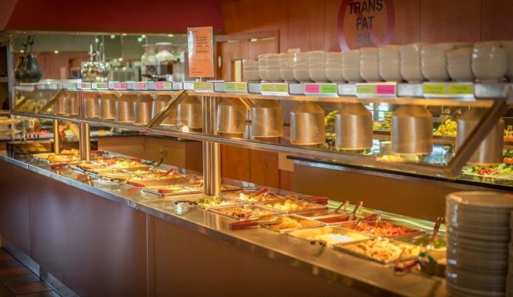 Discover one of the premier buffets in Edmonton, presenting an array of hot and cold dishes featuring fresh vegetables, succulent meats, and crisp salads under inviting warm lighting. With plates stacked to the side for your self-service convenience, it's a culinary experience not to be missed.