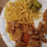 A plate featuring crispy fried chicken strips, savory noodles, and tangy green olives sits enticingly next to a salt shaker, reminiscent of the diverse offerings found at buffets in Edmonton.