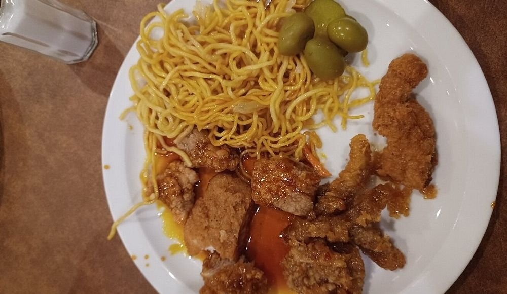 A plate featuring crispy fried chicken strips, savory noodles, and tangy green olives sits enticingly next to a salt shaker, reminiscent of the diverse offerings found at buffets in Edmonton.