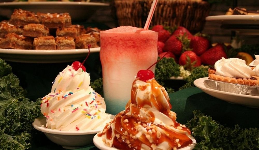Two bowls of ice cream topped with delightful extras, a colorful frozen drink with a straw, and an array of desserts in the background, including strawberries and pies—just like the sweet selection you'd find at the best buffets in Edmonton.
