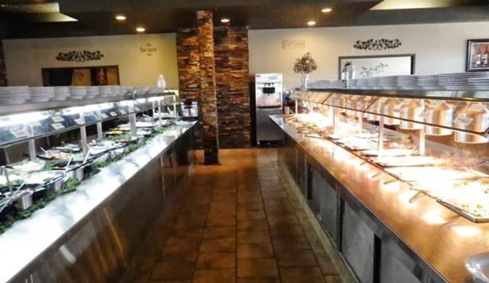 This Edmonton buffet restaurant features two extensive food-serving counters—one brimming with fresh salad options and the other lined with hot dishes. The cozy interior showcases warm lighting and a charming brick column at the end.