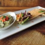 A platter featuring a delightful spinach dip garnished with chopped tomatoes, surrounded by pita bread slices and mixed greens, reminiscent of the diverse offerings found at buffets in Edmonton, rests invitingly on a wooden table.