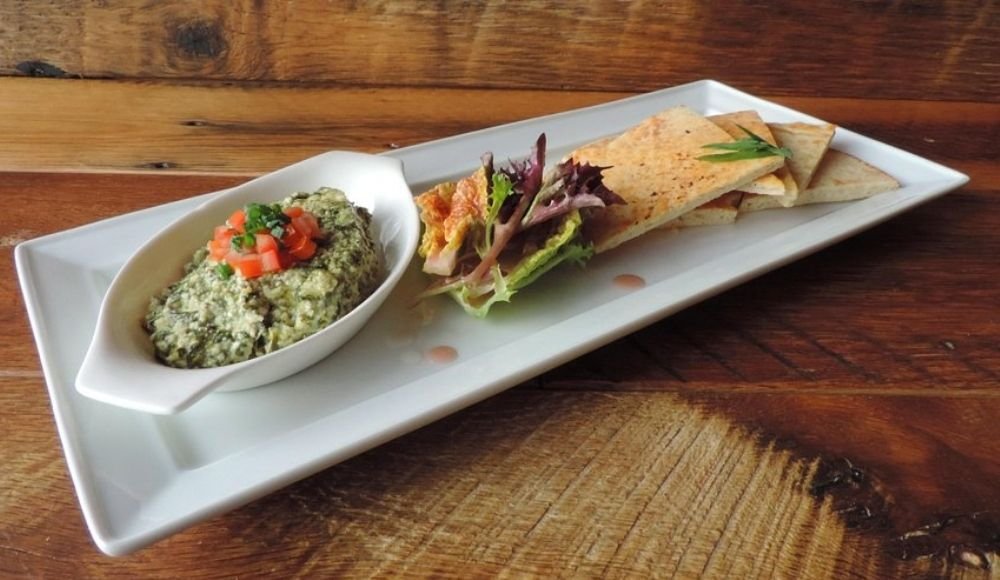A platter featuring a delightful spinach dip garnished with chopped tomatoes, surrounded by pita bread slices and mixed greens, reminiscent of the diverse offerings found at buffets in Edmonton, rests invitingly on a wooden table.