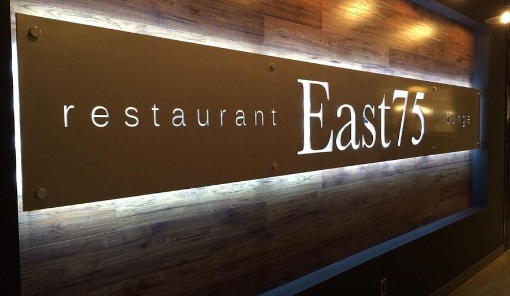 Sign for East75 restaurant and lounge on a wooden wall background, featuring a modern design and illuminated lettering. Known for offering one of the best buffets in Edmonton, it’s a spot where style meets delectable dining experiences.