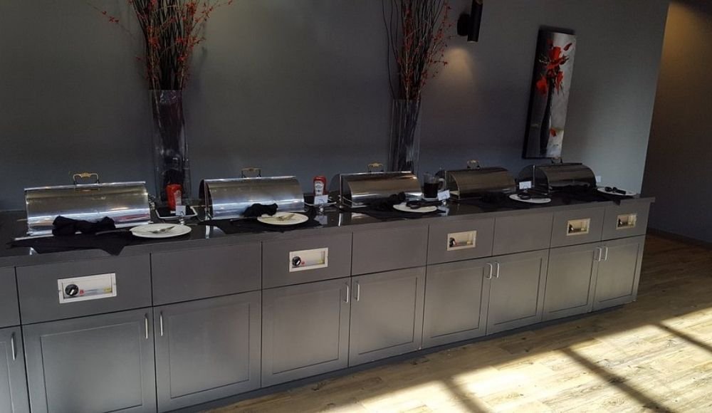 A row of closed chafing dishes lines the countertop against a gray wall, embodying the minimalist style of buffets in Edmonton, all atop a sleek wooden floor.