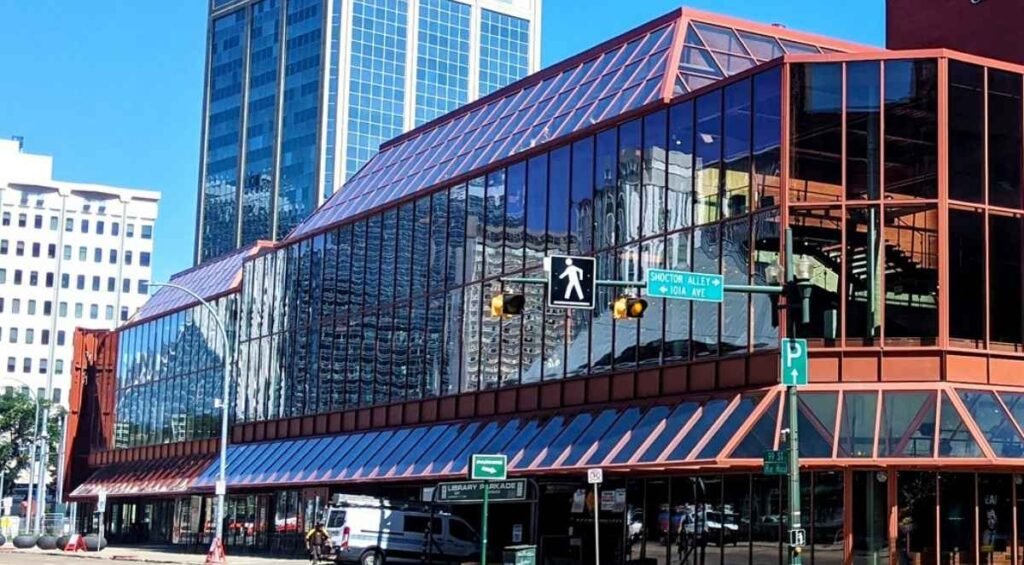 A large building with glass walls reflecting surrounding structures and street signs. Traffic lights and street signs are visible in the foreground, along with a few parked vehicles—one of many modern attractions in Edmonton.